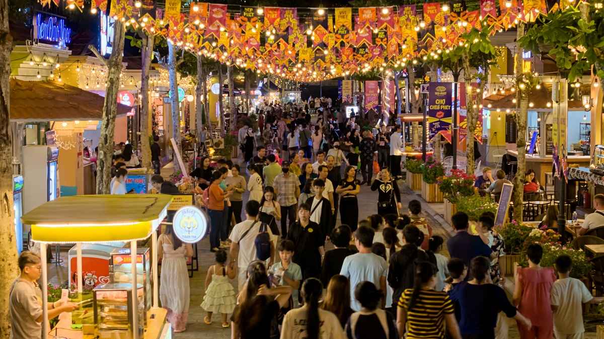 VUI-Fest Bazaar with strings of overhead lights illuminating stalls and a crowd of people near the sea at sunset